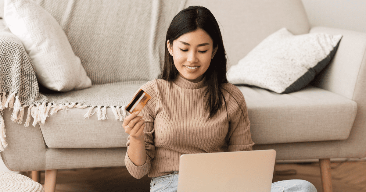 A woman in a beige shirt is sitting on the floor and holding a credit card while looking at her laptop